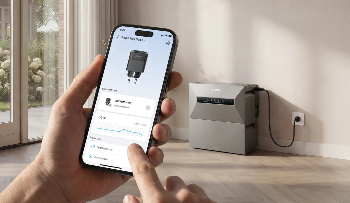 De Anker SOLIX Smart Plug Gen 2 aangesloten in een stopcontact, met op de achtergrond een huishoudelijk apparaat en een smartphone waarop de Anker SOLIX-app realtime het energieverbruik en de energiestromen toont.